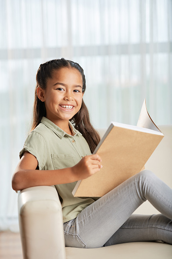 Cute Asian girl smiling and looking at camera while reading good book on sofa